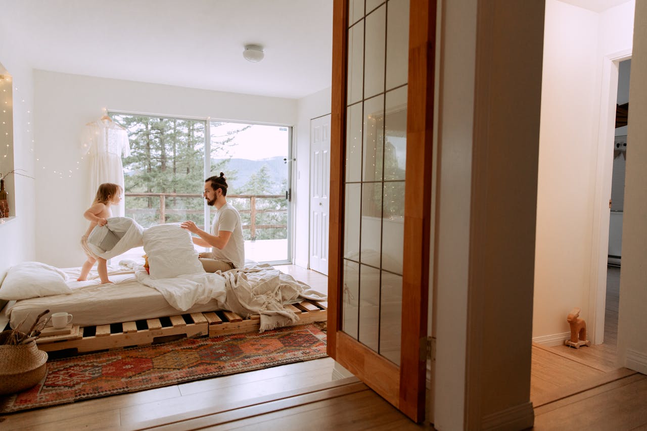 A father and child sharing playful moments in a cozy, light-filled bedroom setting.